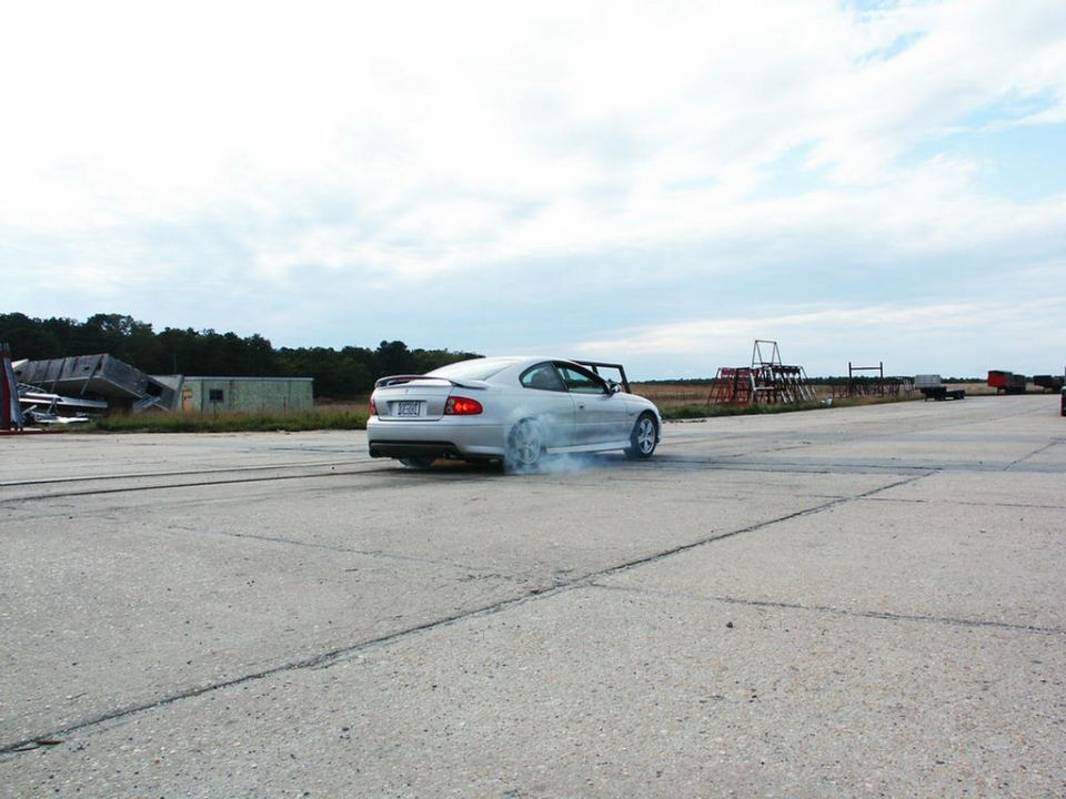 Silver coupe doing a burnout on a paved surface, smoke coming from rear tires. Building debris in background