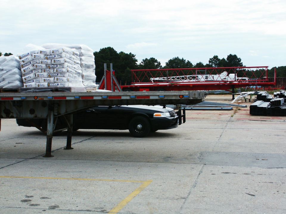 Black sedan, looks like a Crown Victoria, on a flatbed trailer with bags of material and crane parts