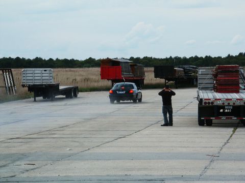 2001 Mercury Grand Marquis - A person stands in a lot as a car drives away, with several flatbed trailers loaded with materials in the background.