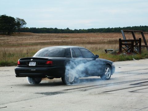 2001 Mercury Grand Marquis - A black sedan performs a burnout, creating a large cloud of smoke on a paved surface with a field in the background.