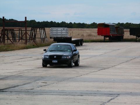 2001 Mercury Grand Marquis - A black sedan is seen from the front on a concrete surface, with trailers and industrial structures in the background.