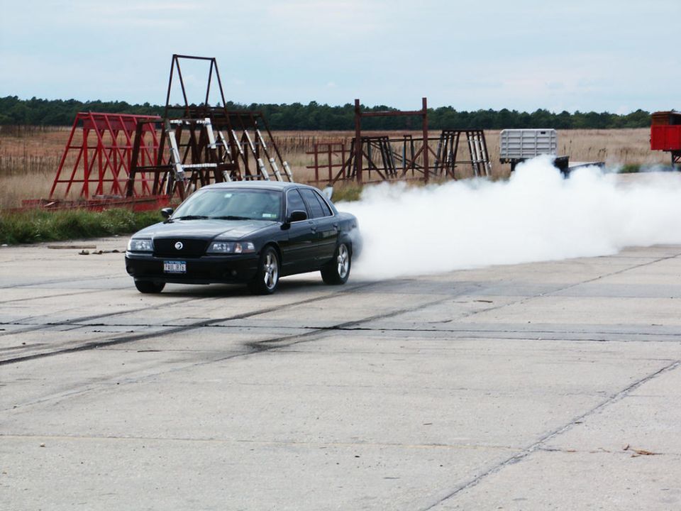 Black sedan with chrome wheels doing a burnout, emitting white smoke. License plate visible