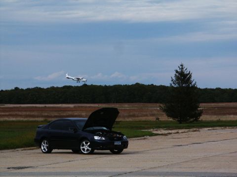 2001 Mercury Grand Marquis - Dark sedan with hood open parked on pavement, small twin-engine airplane flying in the sky.