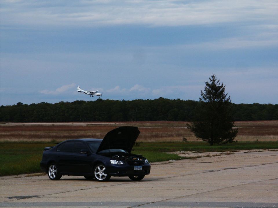 Dark sedan with hood open parked on pavement, small twin-engine airplane flying in the sky