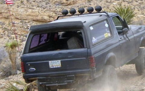 1987 Ford Bronco - A dark gray Ford Bronco navigates a dusty off-road trail with a spare tire visible in the rear.