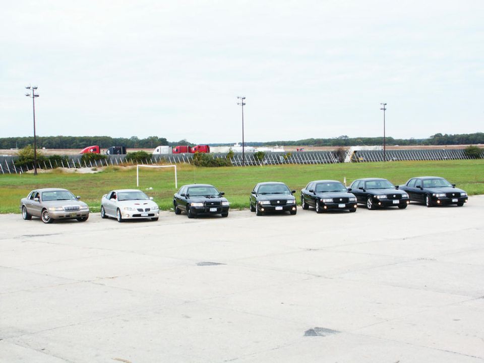 Seven sedans parked in a row on pavement, with trucks and a field in the background