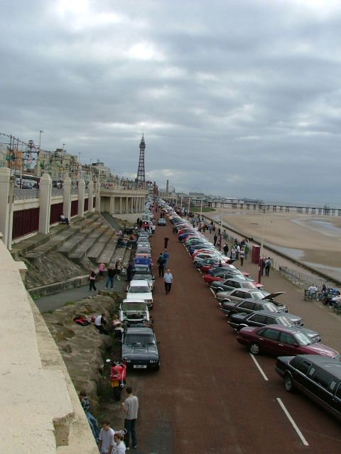 2001 Ford Fiesta - A long line of vehicles are parked on a promenade next to a beach, with a tall tower visible in the distance.