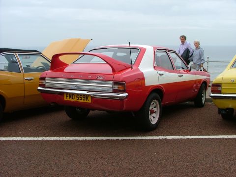 2001 Ford Fiesta - A red and white Ford Cortina is parked next to other vehicles, with two people standing in the background.