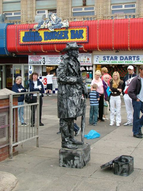 2001 Ford Fiesta - A silver-painted street performer poses in front of a burger bar and arcade on a sunny day.