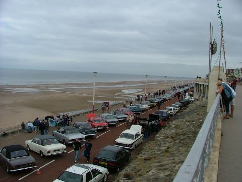 2001 Ford Fiesta - A long line of classic cars are parked along a promenade next to a beach under a cloudy sky.