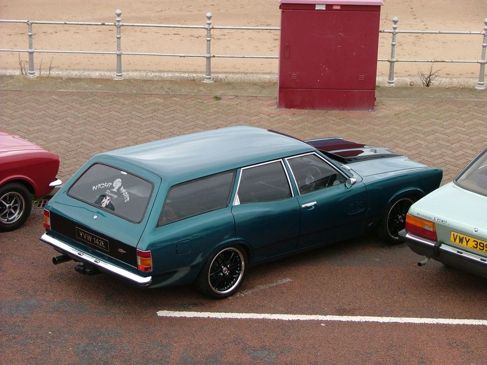Teal station wagon parked next to a red car and a light blue car. Aftermarket wheels and a hood scoop are visible