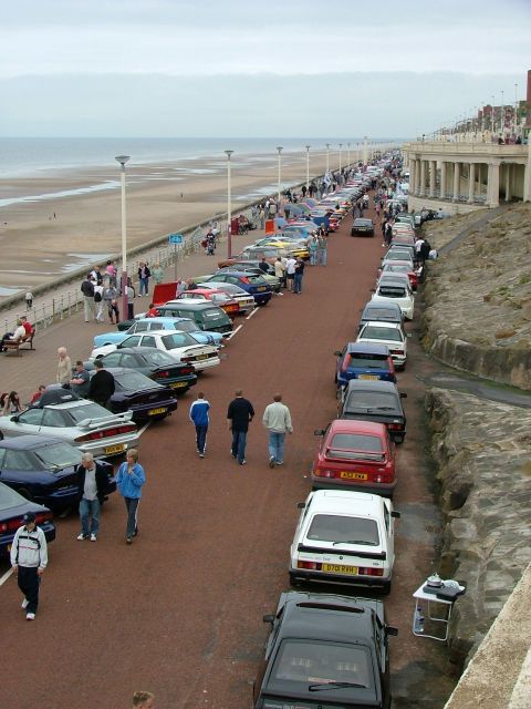 2001 Ford Fiesta - A long line of vehicles are parked on a paved area next to a beach and a building with columns.