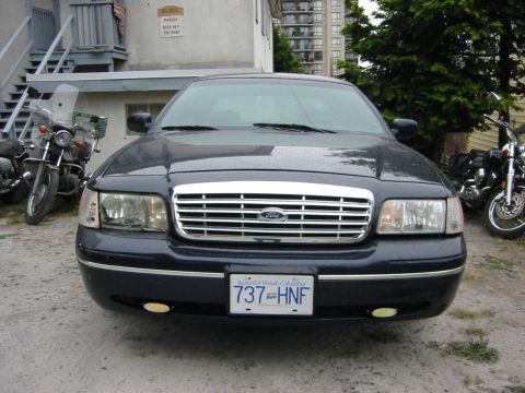 1999 Ford Crown Victoria - A dark blue Ford Crown Victoria is parked outside a building with two motorcycles nearby.