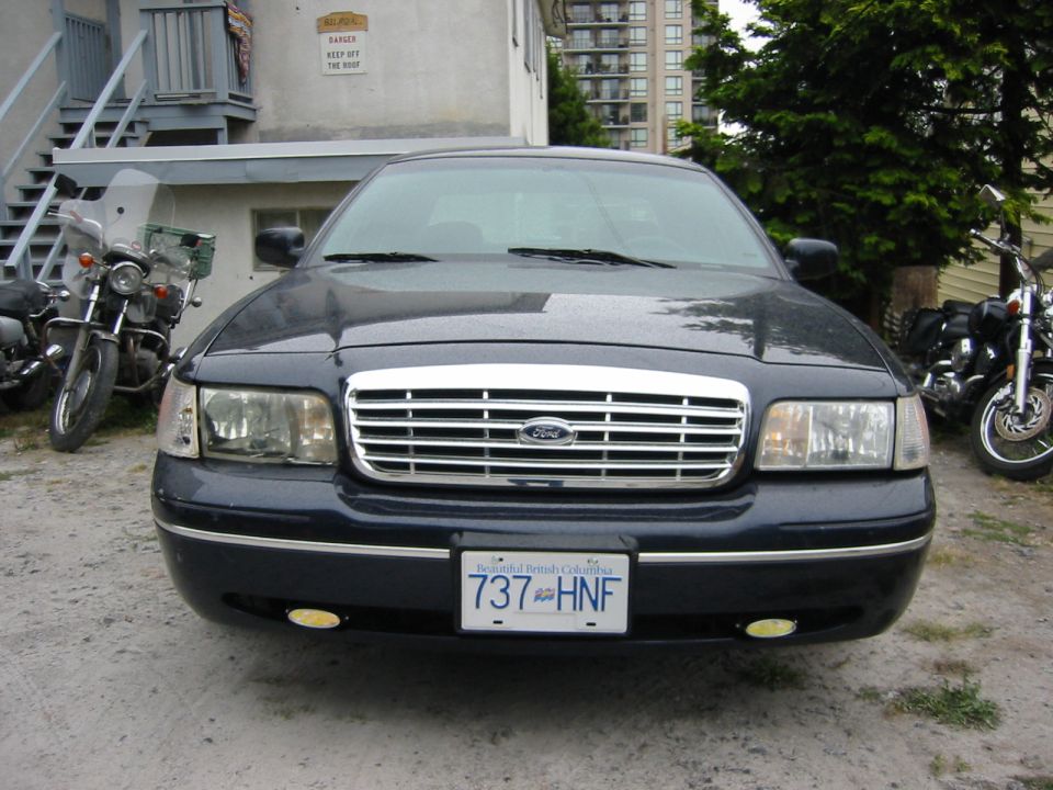 Front view of a dark blue Ford Crown Victoria with a chrome grille. Two motorcycles are parked on either side