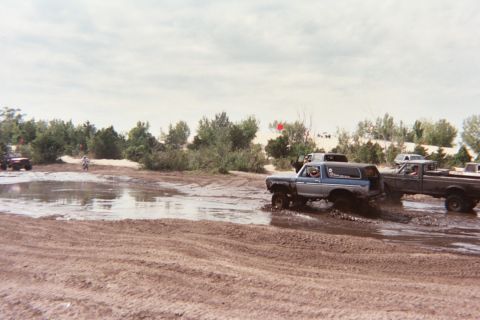 1979 Ford Bronco - Off-road vehicles navigate a muddy terrain with trees and a cloudy sky in the background.