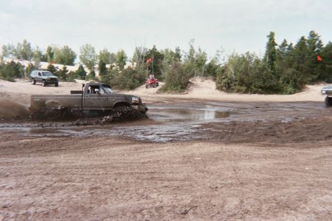 1979 Ford Bronco - A pickup truck navigates a muddy off-road course, kicking up water as an SUV and ATV are visible in the distance.