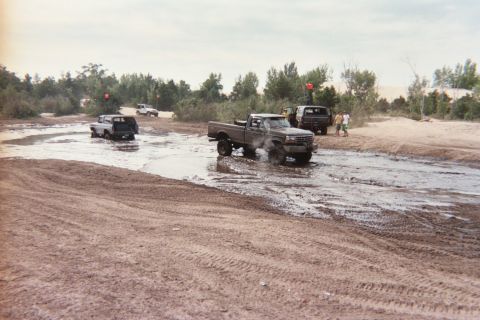 1979 Ford Bronco - Multiple trucks navigate a muddy terrain, with spectators observing the off-road activity.