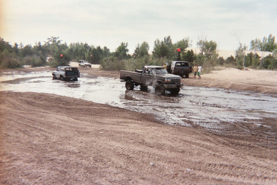Several trucks driving through a muddy area with trees and sand dunes in the background