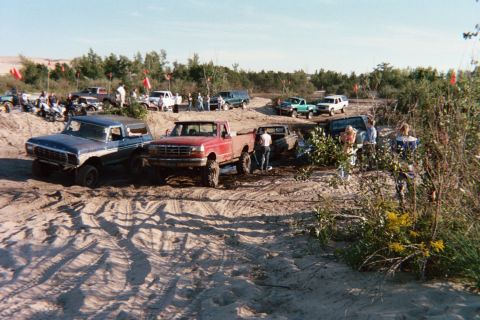 1979 Ford Bronco - Several off-road vehicles are gathered in a sandy area with trees and people around.