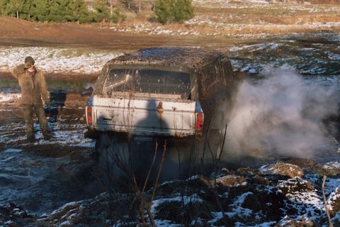 1979 Ford Bronco - A person watches as a muddy SUV struggles to get out of a water-filled ditch.
