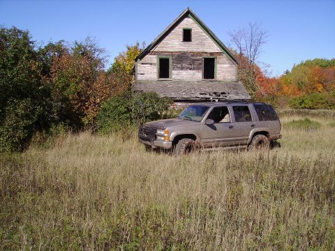 2000 Chevrolet Tahoe - A dirty Chevrolet Tahoe sits in a field of tall grass before a dilapidated house under a clear blue sky.