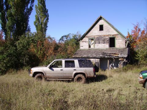 2000 Chevrolet Tahoe - A silver SUV sits in a field of tall grass, with an old, weathered house in the background. Another vehicle is visible to the right.