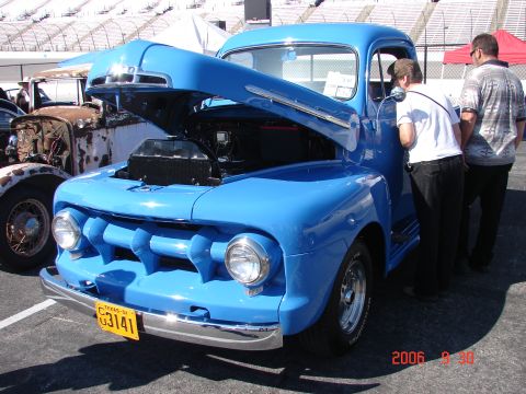 1987 Ford Bronco - A bright blue pickup truck is displayed with its hood open, while two people look on. Another older truck is visible in the background.