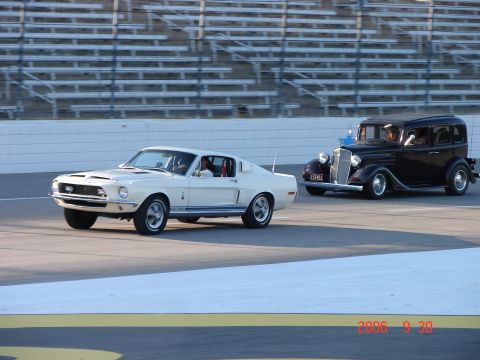 1987 Ford Bronco - Two classic cars race on a track with grandstands in the background.