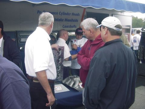 1990 Ford Mustang - People examine engine parts at an outdoor event, with a sign advertising performance parts in the background.