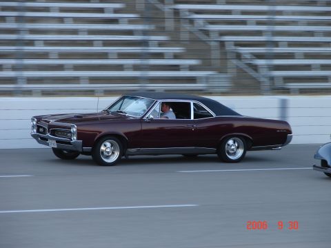 1987 Ford Bronco - Maroon 1966 Pontiac GTO coupe with a black vinyl top driving on a track, with a partial view of another vehicle to the right.