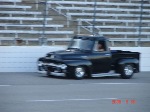 1987 Ford Bronco - A black 1956 Ford F-100 pickup truck drives on a race track, with bleachers in the background.
