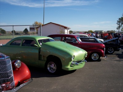 1972 Ford Ranchero - A line of cars are parked outdoors under a clear blue sky, with a prominent green hot rod in the foreground.