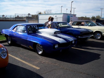 1972 Ford Ranchero - A blue 1969 Camaro drag car with racing numbers is parked in a lot with other vehicles under a partly cloudy sky.