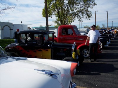 1972 Ford Ranchero - Several custom vehicles are parked in a lot under a bright sky, with a man standing next to a red pickup.