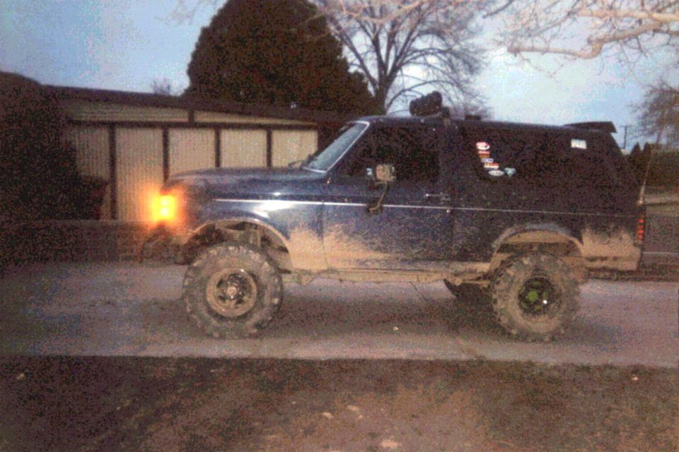 Lifted dark blue Ford Bronco on mud tires at dusk, mud-splattered rockers, gravel lot