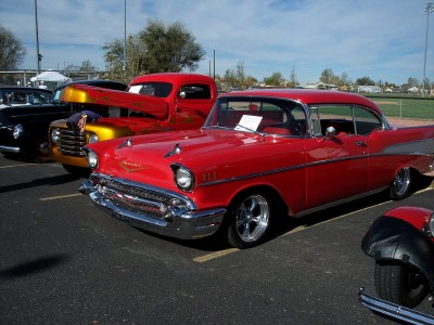 1972 Ford Ranchero - A row of classic vehicles, including a red 1957 Chevrolet and a red pickup truck, are parked outdoors on asphalt under a bright blue sky.