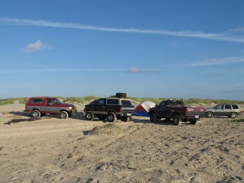 1995 Ford Bronco - Four four-wheel-drive vehicles lined up on a sandy beach under a blue sky, a red-and-silver Ford Bronco, a black Bronco with a roof-rack sp…
