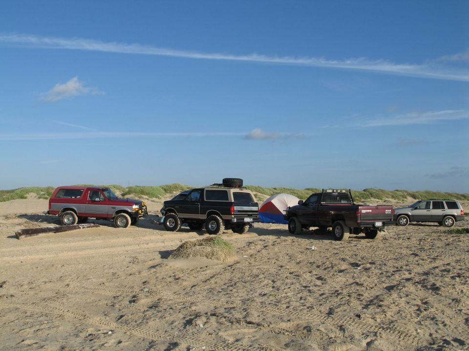 Four 4x4s lined up on a beach: two Ford Broncos, a Dodge pickup and a Jeep, with a tent