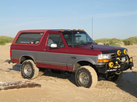 1995 Ford Bronco - Lifted two-tone red-over-silver full-size Bronco parked on sand dunes, shown from the 3/4 front-passenger side on knobby mud tires with whi…