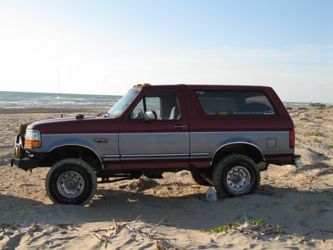 1995 Ford Bronco - Two-tone maroon over silver full-size Ford Bronco viewed from the driver's side, parked on a sandy beach with ocean surf behind it. It is l…
