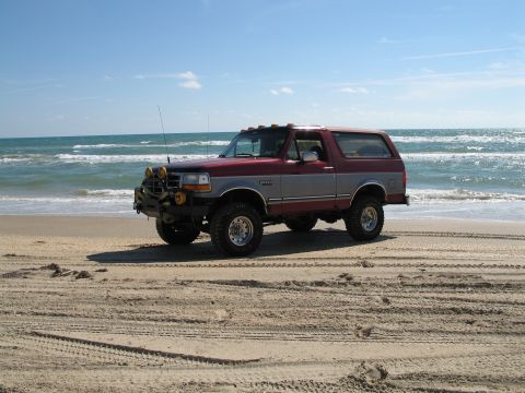1995 Ford Bronco - A two-tone maroon-and-silver full-size Ford Bronco with a hardtop, lifted on mud tires, parked on a sandy beach with the ocean behind; fron…