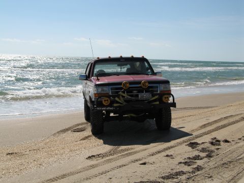 1995 Ford Bronco - Lifted red/maroon Ford Bronco parked on wet sand at the surf line, viewed head-on with tire tracks running down the beach. It runs a black…