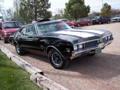 1972 Ford Ranchero - A black muscle car with white racing stripes is parked on a gravel lot, with other vehicles visible in the background.