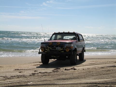 1995 Ford Bronco - A lifted 1990s full-size Ford Bronco in faded red parked on a sandy beach with the ocean breaking behind it. It wears a front brush guard,…