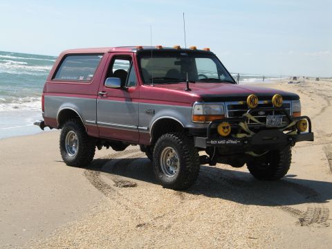 1995 Ford Bronco - Red and silver two-tone full-size Ford Bronco parked on a beach at the surf line, shot from the front three-quarter. It's lifted on mud-ter…