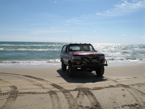 1995 Ford Bronco - Maroon full-size Ford Bronco parked at the water's edge on a sandy beach, seen nearly head-on, lifted on mud tires with roof-mounted KC lig…