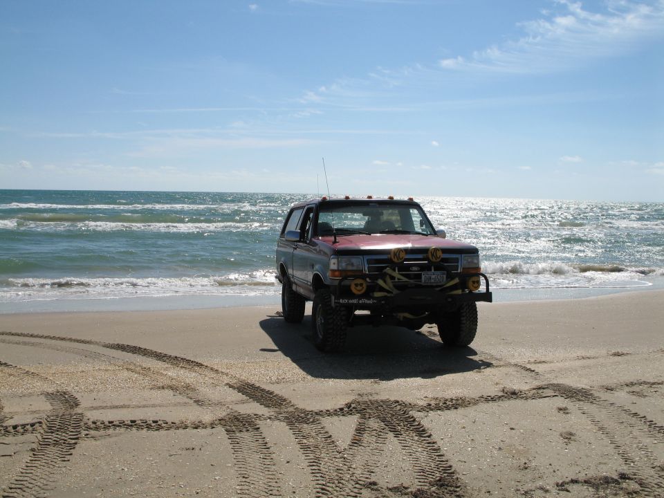 Maroon lifted Ford Bronco with KC lights parked on a beach by the surf