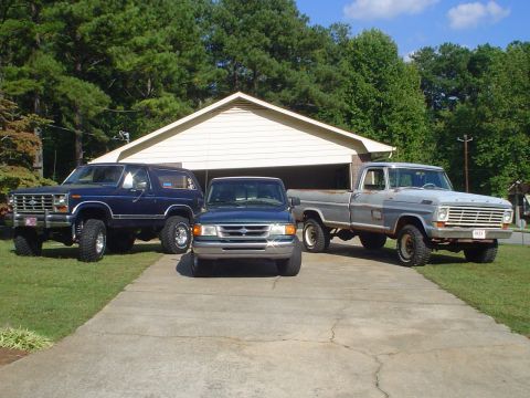 1984 Ford Bronco - Three Ford trucks and SUVs are parked in front of a garage on a sunny day.