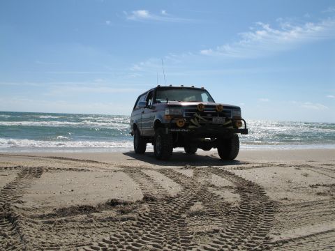 1995 Ford Bronco - A lifted Ford Bronco parked on a beach at the surf line, wearing a tubular front bumper with yellow-covered KC driving lights, cab-roof cle…