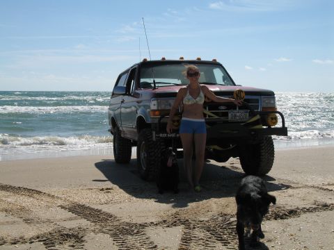 1995 Ford Bronco - A lifted red 1990s full-size Ford Bronco parked on wet sand at the ocean's edge, fitted with a tube brush guard and winch, yellow KC drivin…